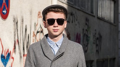 Junger Mann mit dunkler Sonnenbrille, grauem Mantel und Schiebermütze