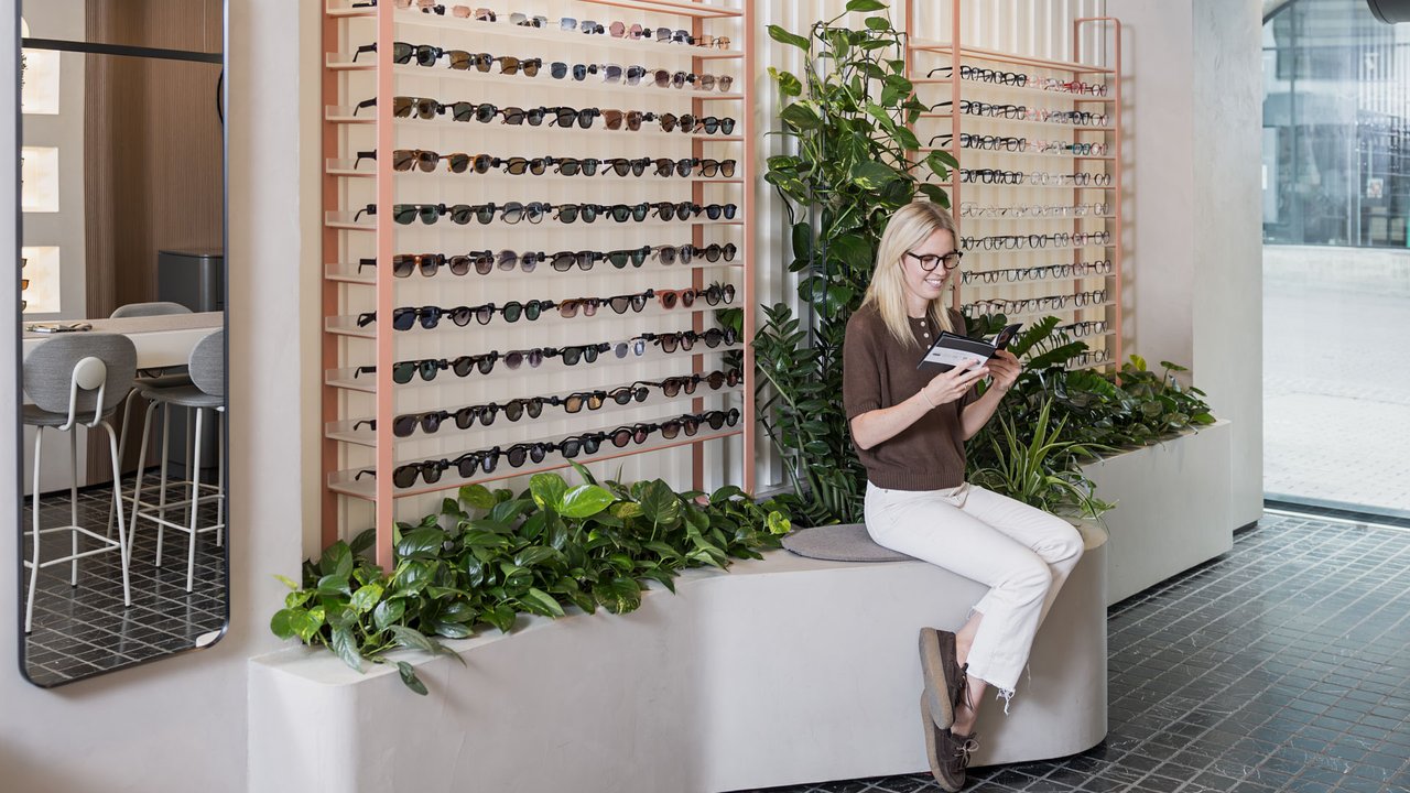 Kundin betrachtet das Brillensortiment in einem modernen Optikgeschäft mit großer Auswahl an Brillen und Sonnenbrillen. Sie sieht dabei in eine Imagebroschüre des Optikers. 